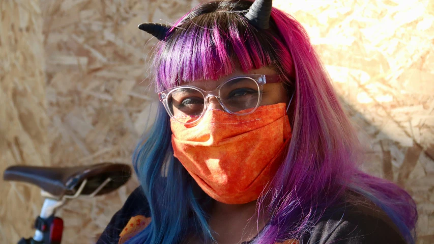 an adult woman with bright colored hair wears a devil mask