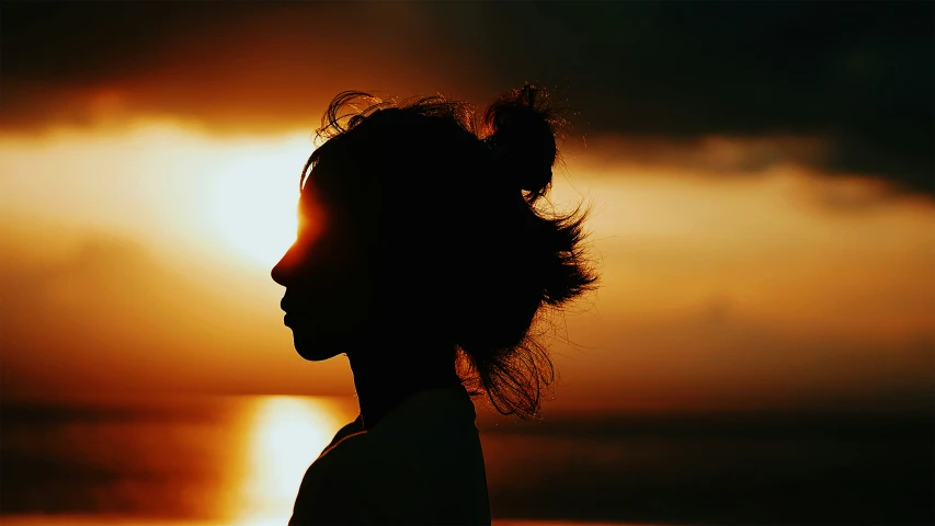 the silhouette of a woman with a messy ponytail, in front of the sunset