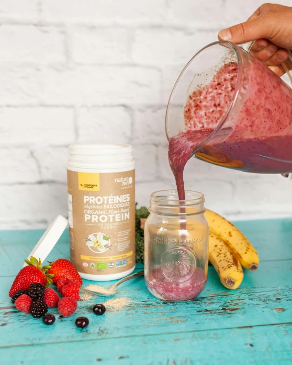 a hand pouring protein into a jar next to bananas, berries, and other ingredients