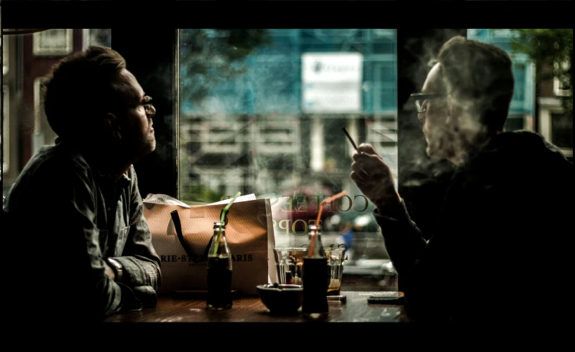 two men sitting at a table in a bar smoking a cigarette