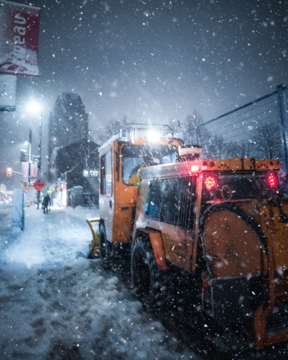 an orange street plow has its headlights on in the snow