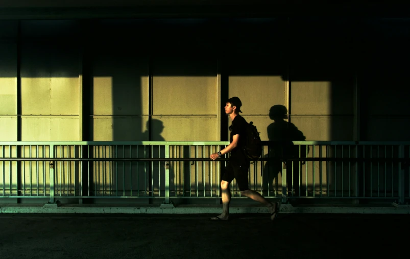 a man walks in a building's shadow near some metal fencing