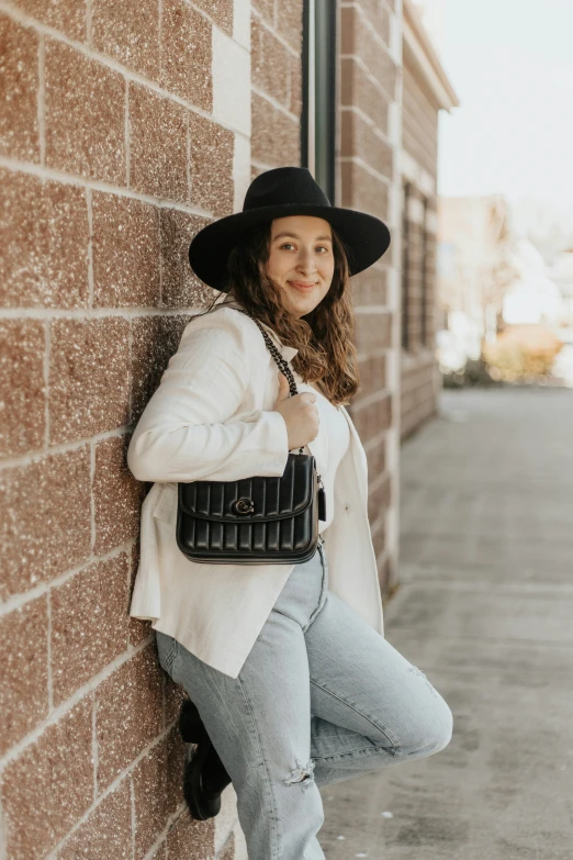 a woman leaning against a brick wall while wearing a black hat