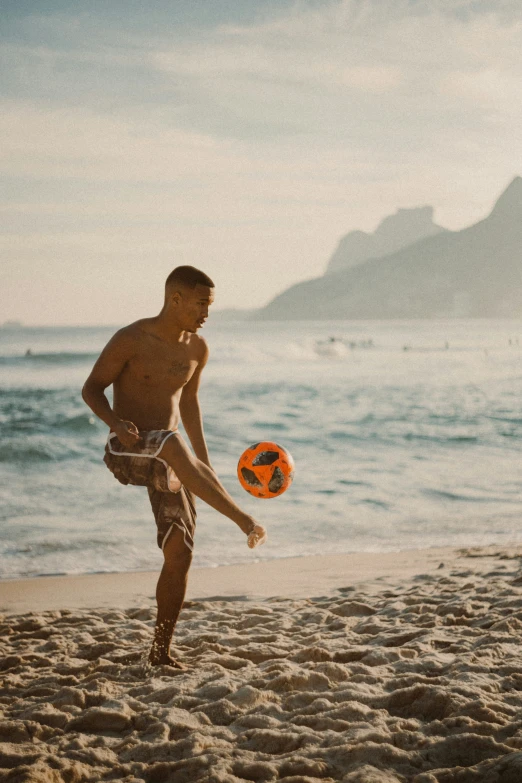 a man in trunks playing with a orange soccer ball on a sandy beach