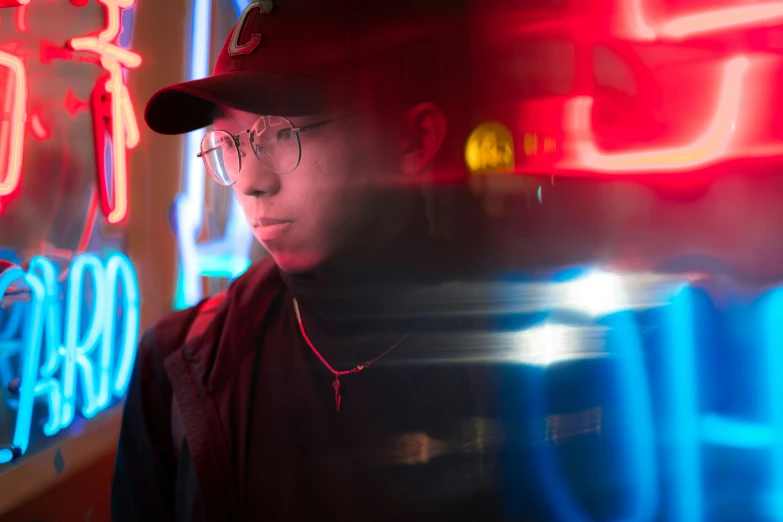 a man in a hat and glasses standing in front of a neon sign