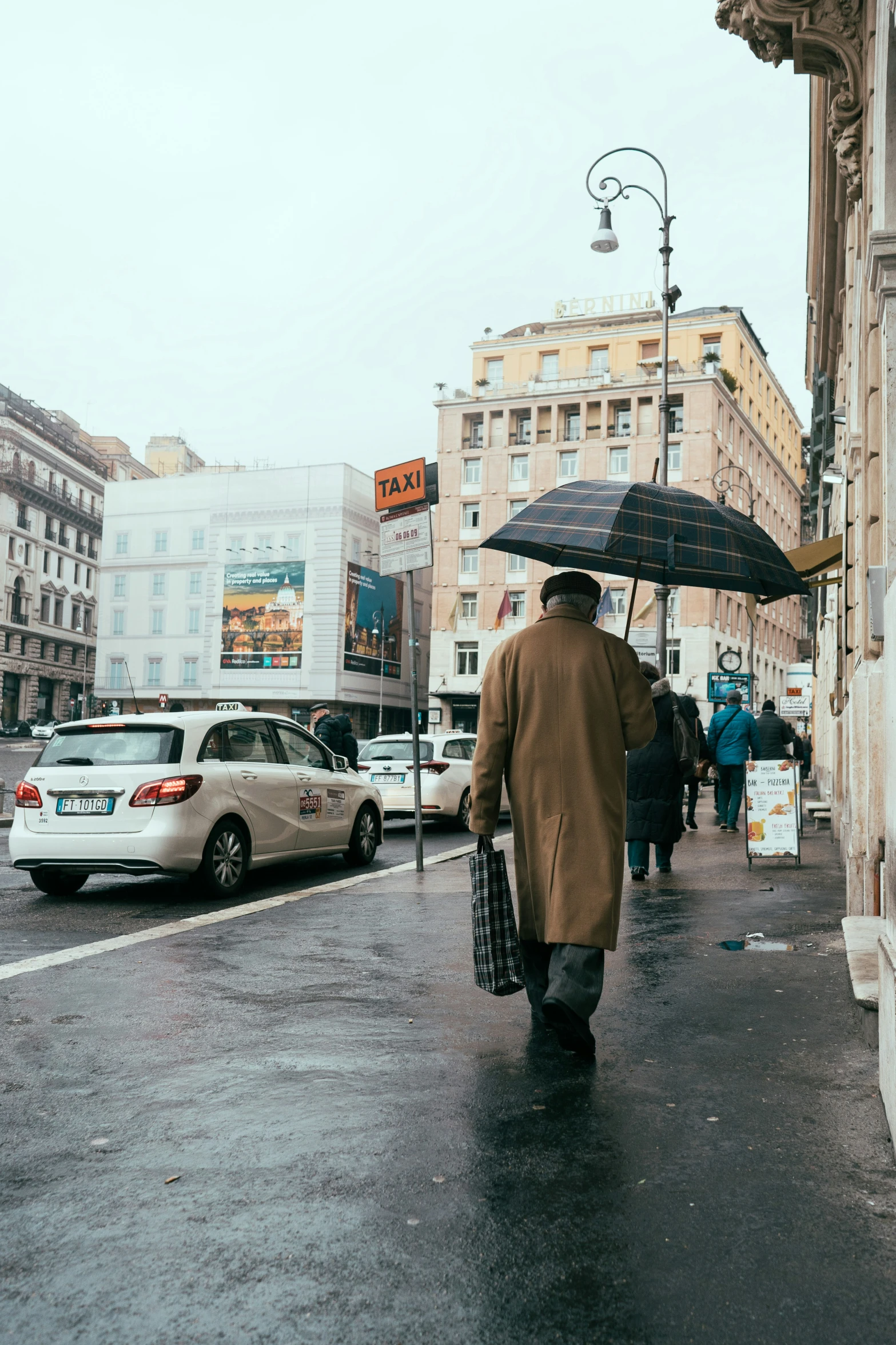 an old man in a brown coat with an umbrella walking on the street