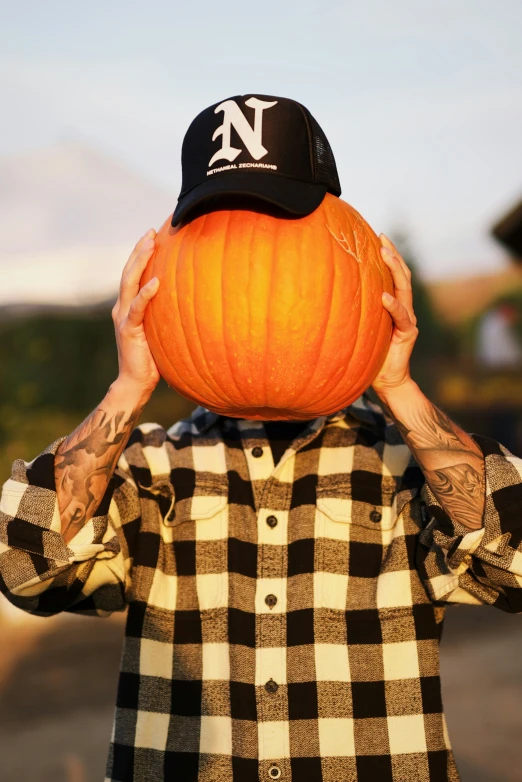 there is a man wearing a ball cap and holding a pumpkin in front of his face