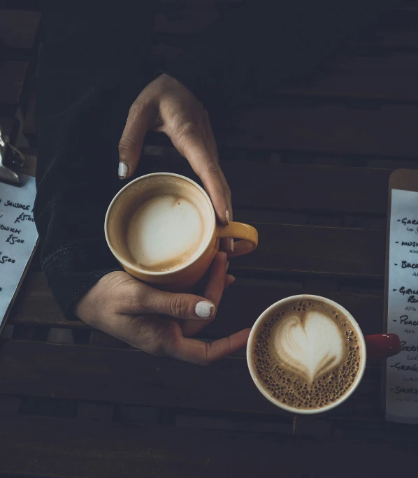 two hands holding cups of coffee on top of a wooden table