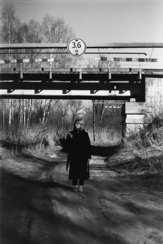 a woman walking down a dirt road under a train bridge
