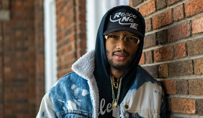 a man with glasses and a hooded sweatshirt leaning against a brick wall