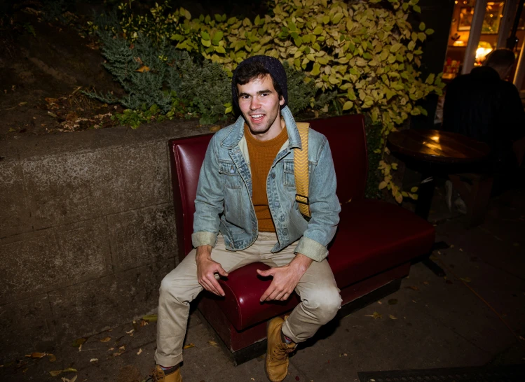 a young man in jeans and a jean jacket smiles while sitting in a red leather chair