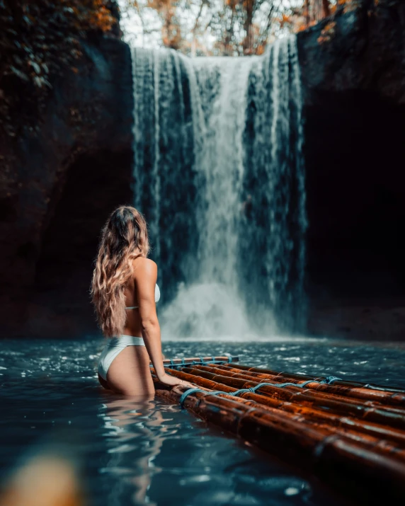 a beautiful woman sitting on top of a wooden raft in front of a waterfall