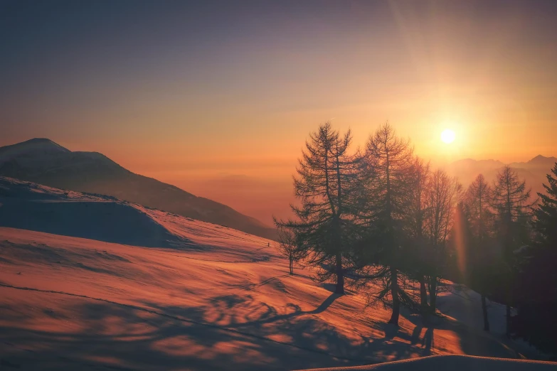 the sun sets behind the trees on top of a snowy mountain