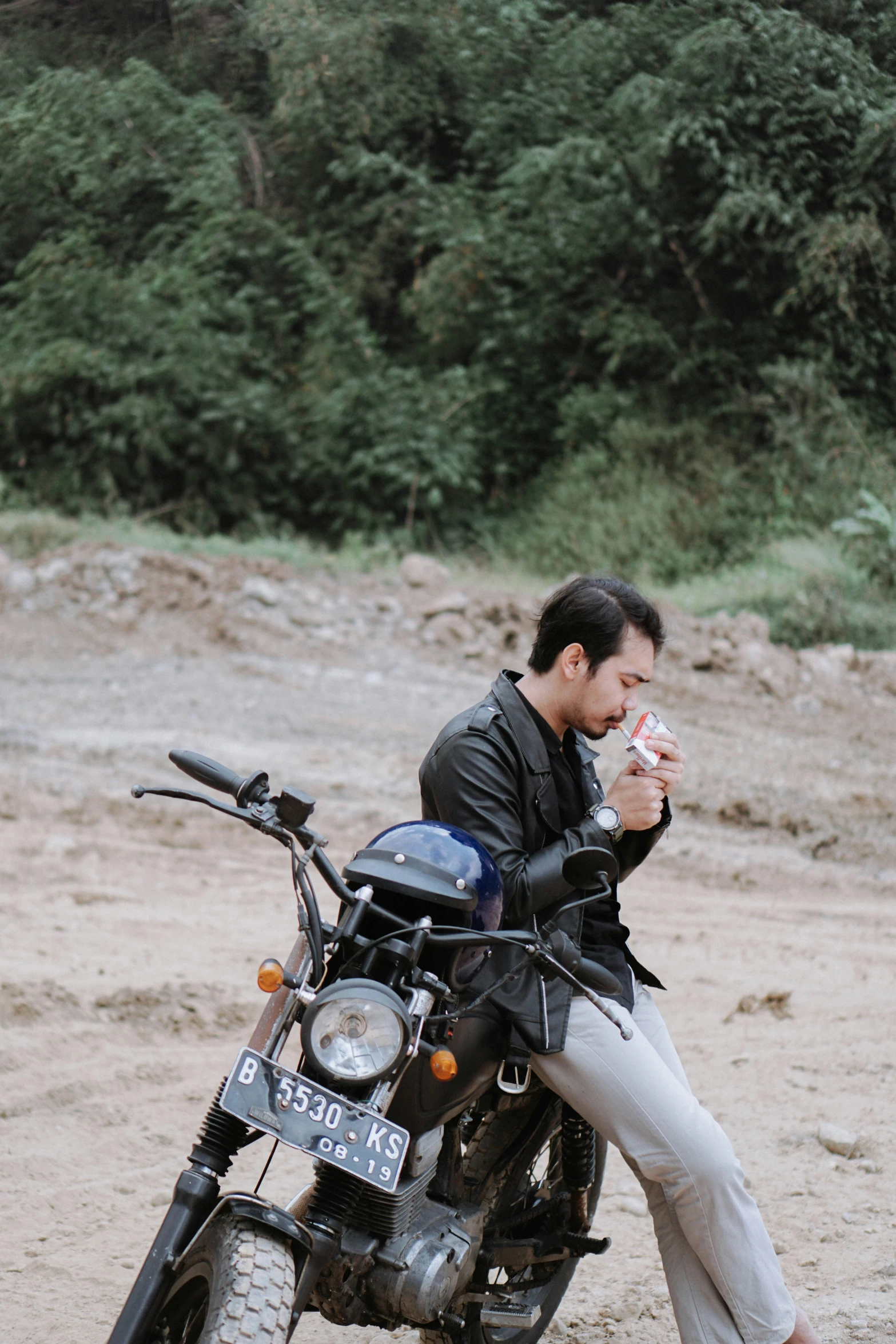 man wearing black jacket and white pants leaning on motorcycle eating ice cream