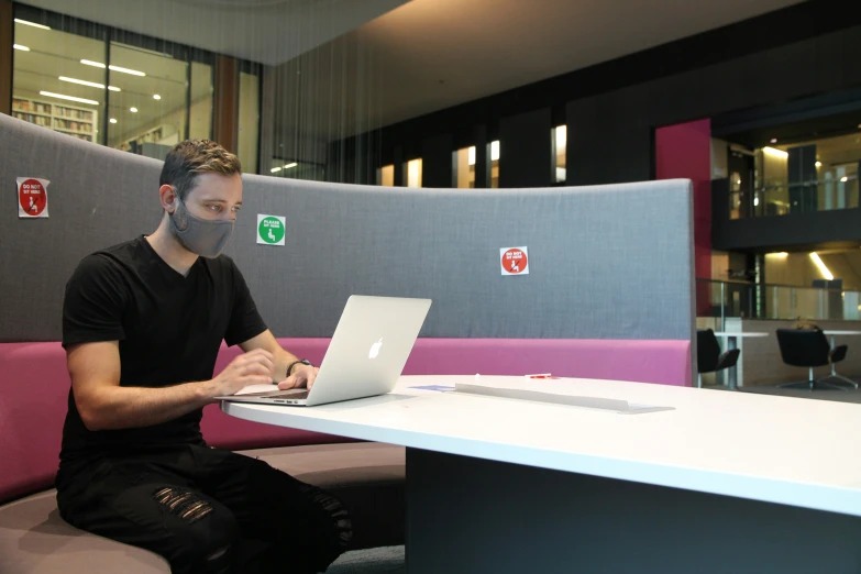 a man with a face mask sitting at a table on a laptop