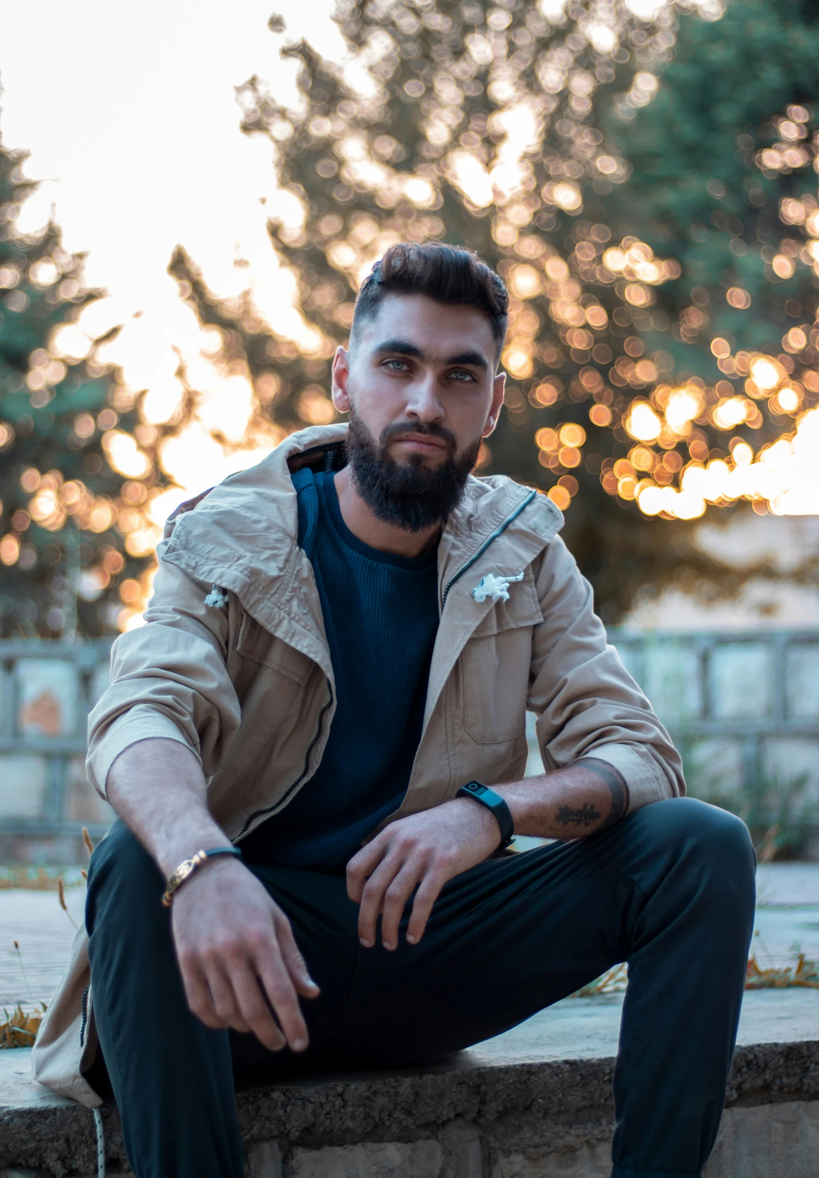 a bearded man sitting on the edge of a wall in front of trees