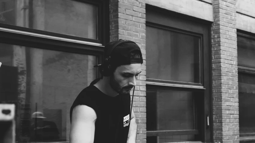 man in black tank top leaning against a building and listening to earbuds