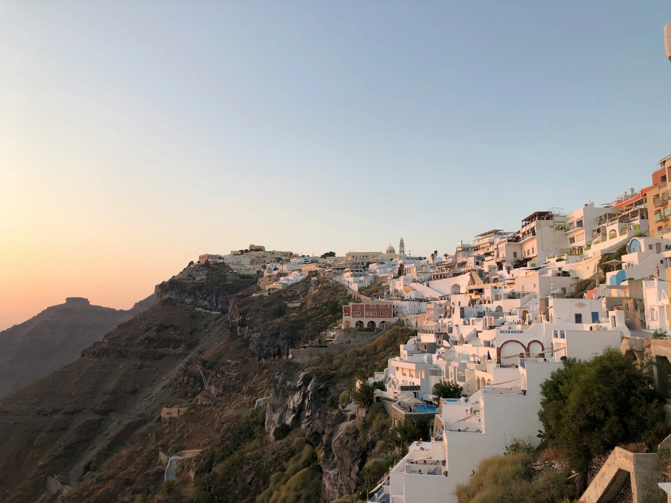 a hillside covered in white colored buildings, with a sunset and ocean in the background