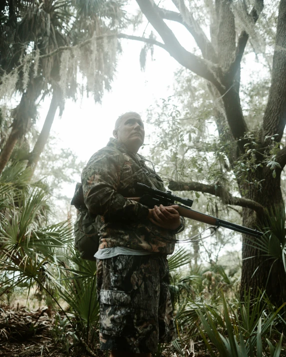 man standing in forest holding rifle with trees and sun in background