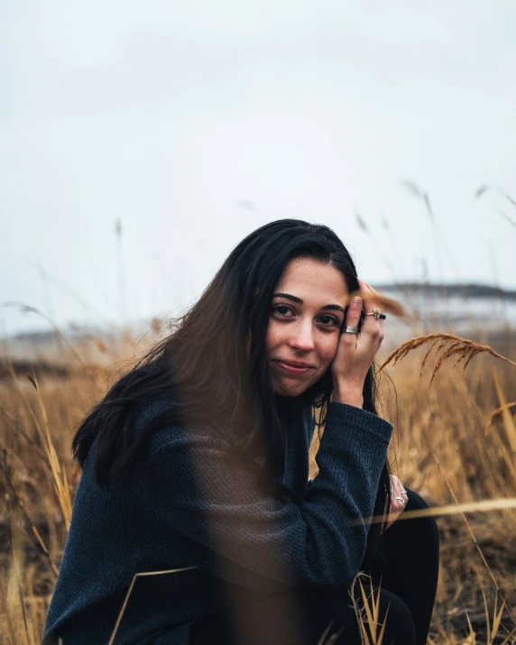 woman sits in tall grass, smiling and holding her ear