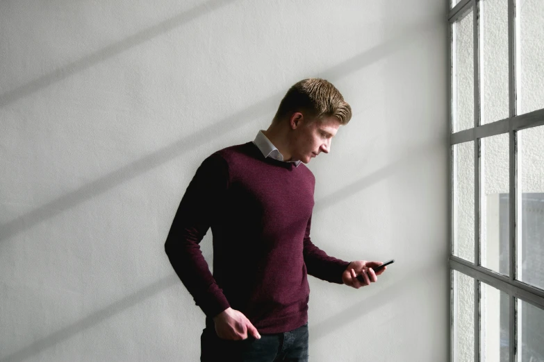 the man is standing next to a window with his cellphone in hand