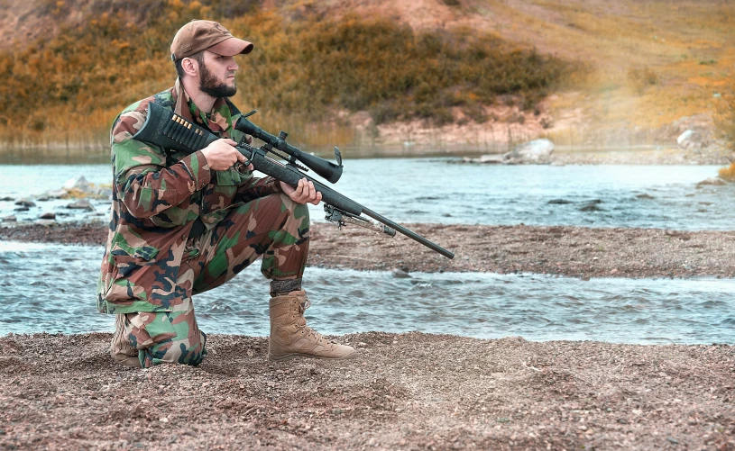 a man squats and holds a rifle and looks at the ground near some water