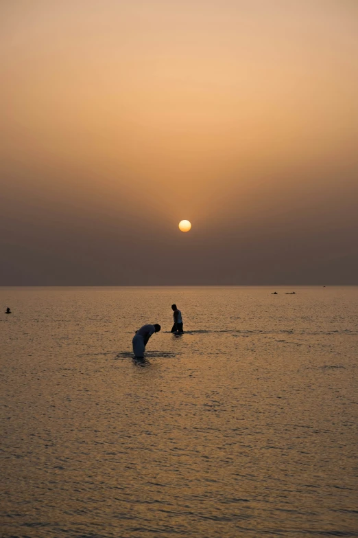 two people sit in the water in front of an orange and yellow sunset