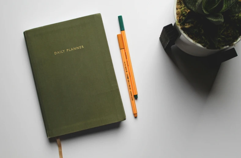 a green notebook with an orange pen and a plant