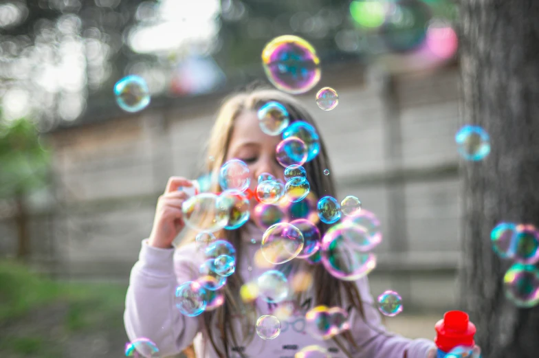 a little girl that is standing by a tree blowing bubbles