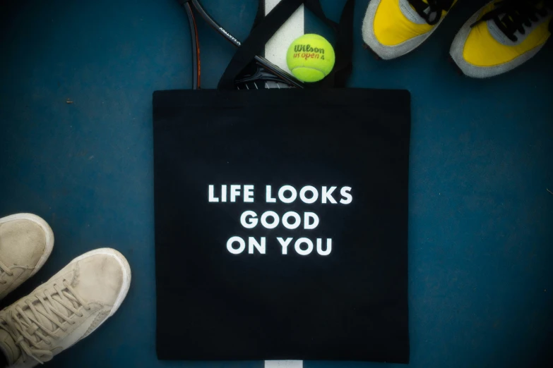 black tote bag that reads life looks good on you, with tennis rackets and ball in background