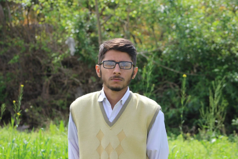 a person in a sweater vest with glasses standing in front of bushes