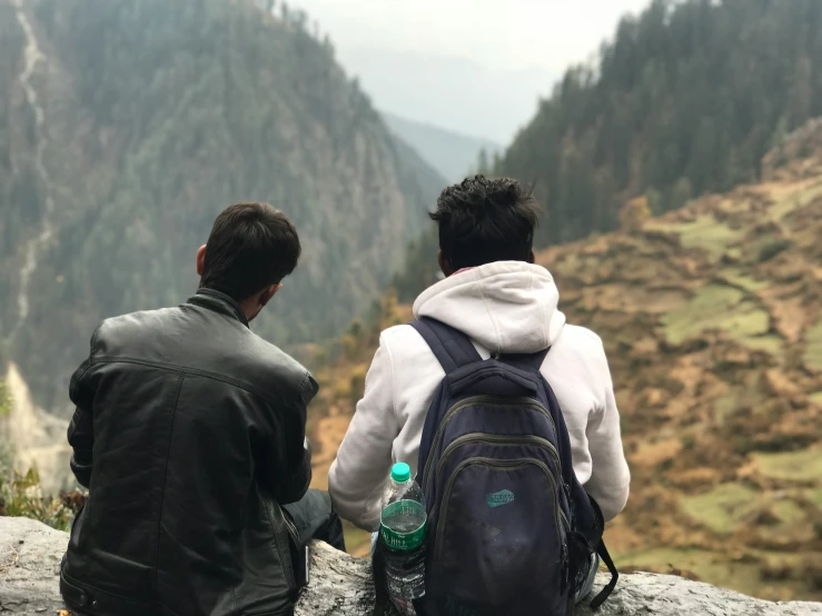 two men sitting together with backpacks facing mountains