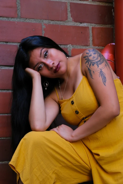 a pretty young lady in a yellow dress leaning against a brick wall