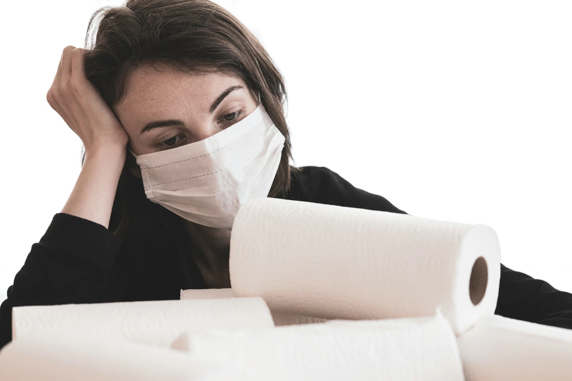 a woman wearing a face mask sitting by several rolls of toilet paper