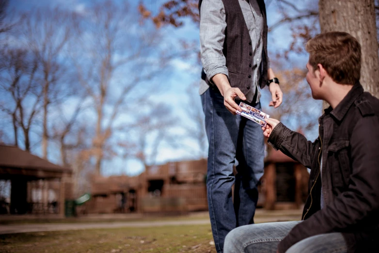 a young man sits by a tree, handing soing to another guy