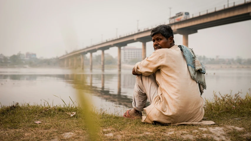 man in a white blanket sitting by a lake near a bridge