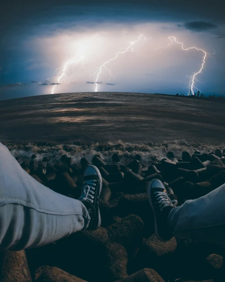 two people in sneakers are watching lightning over the ocean
