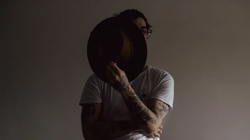 a man with glasses and tattooed arms holding his hat to his face