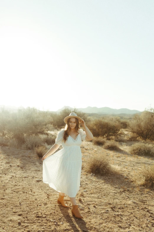 a woman in a white dress and hat walks on a desert path