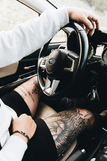 tattooed man driving a car and the driver's feet are visible in his pants