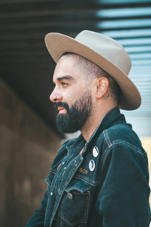 a bearded man wearing a gray hat standing by a wall