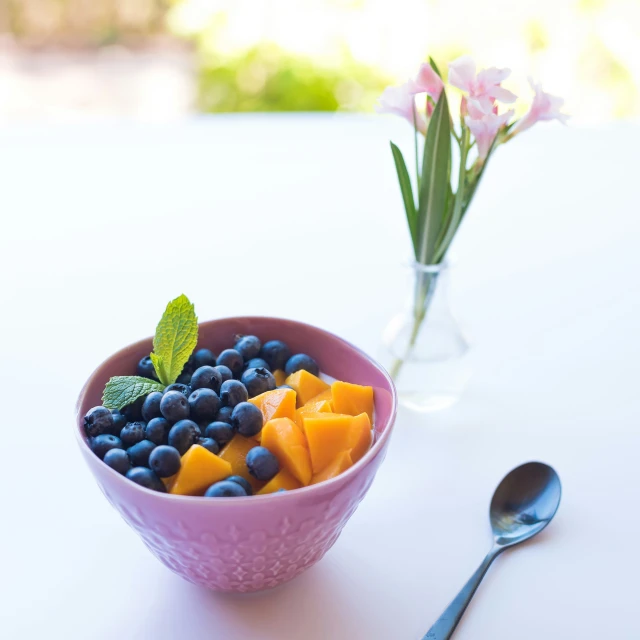 a pink bowl with berries, mango, and blueberries in it next to a glass of water