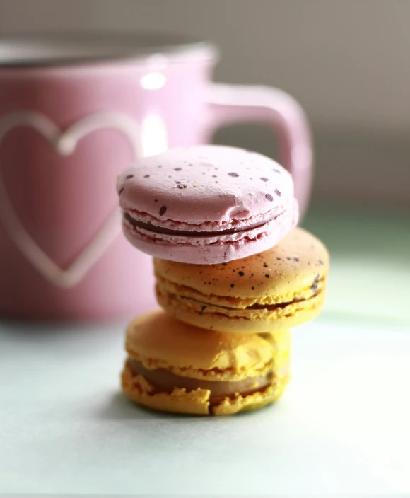 a stack of pink macarons with a heart in a mug behind them