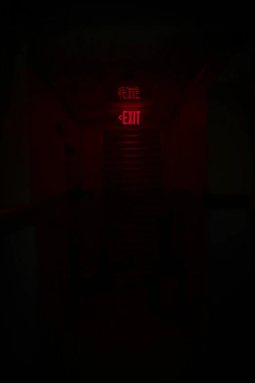 a dark hallway has a red exit light and a creepy staircase