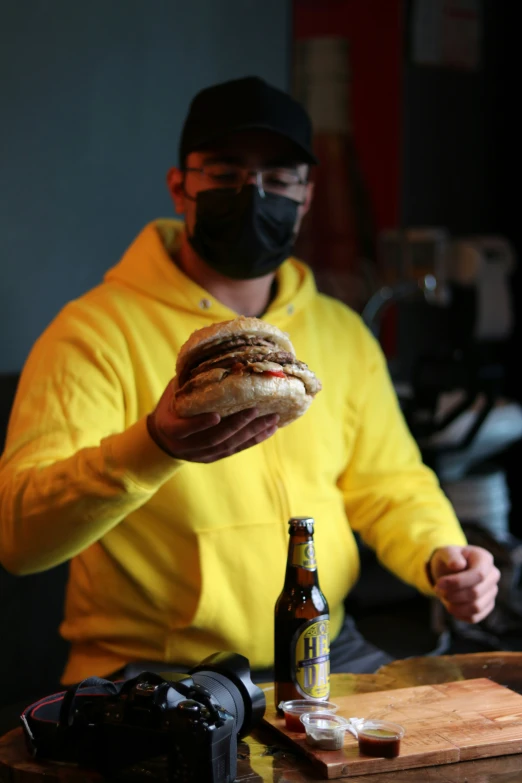 a man is wearing a face mask while holding a large sandwich and a bottle of beer