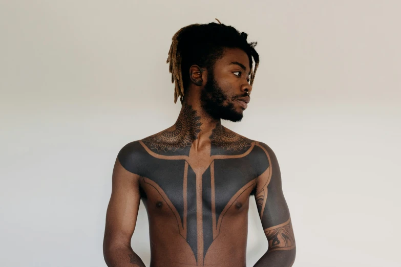 a black man with tattoos on his chest standing against a wall
