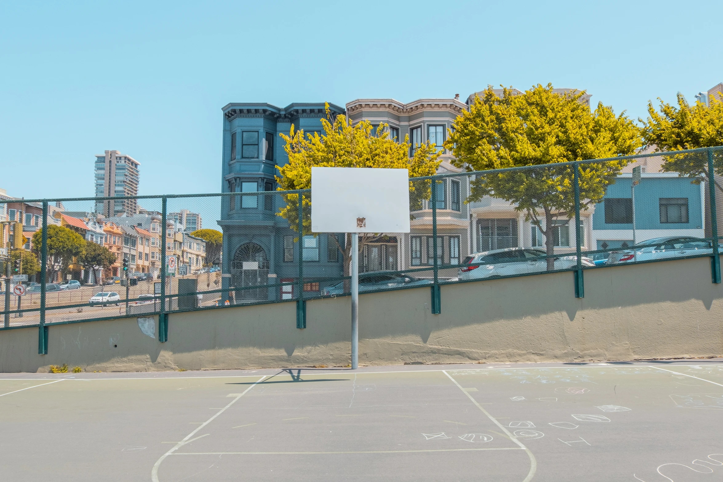 an empty basketball court and basketball hoop next to a fence with buildings in the background