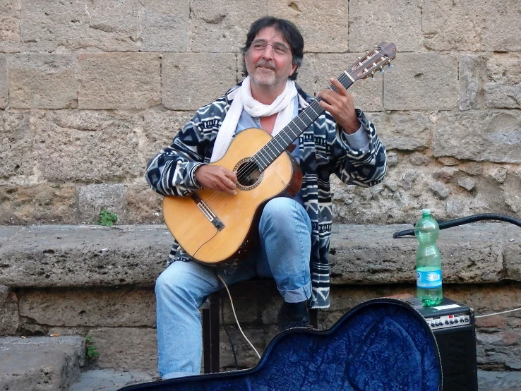 man in patterned jacket sitting and playing guitar next to microphone and guitar case