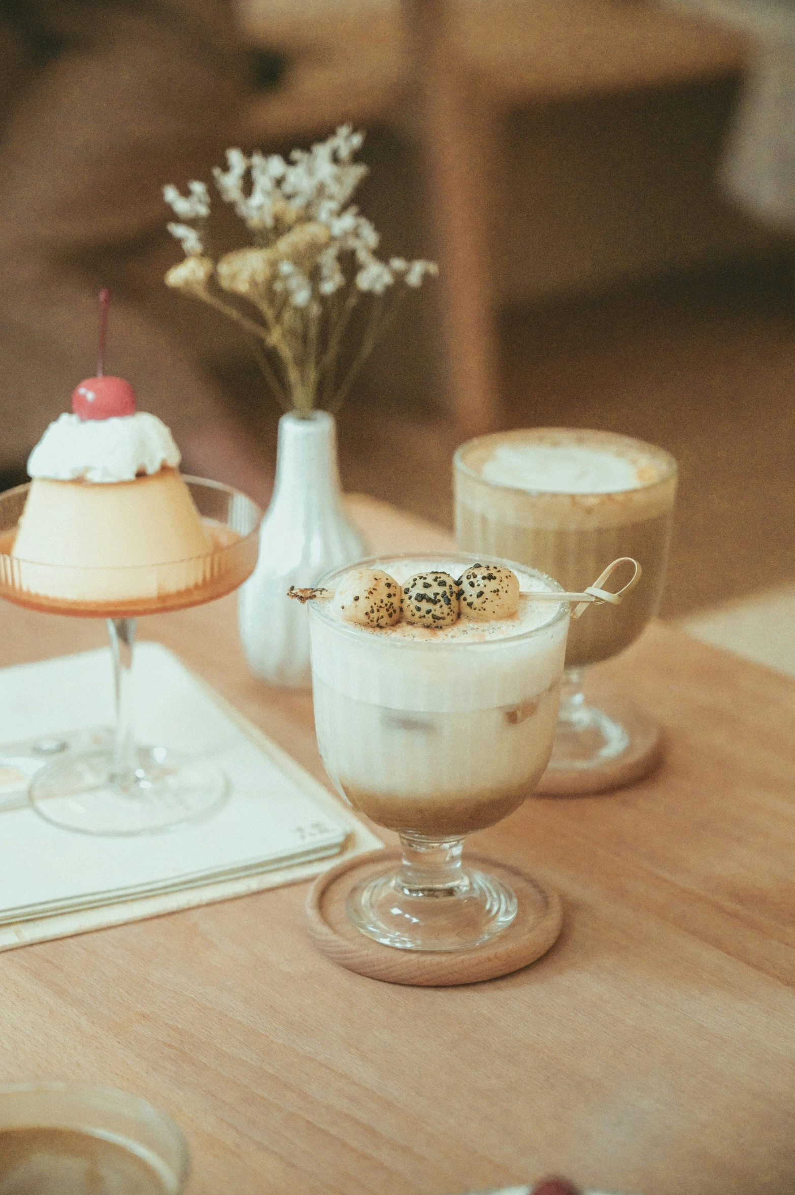 two glasses on a table with a dessert and flowers in a vase behind them