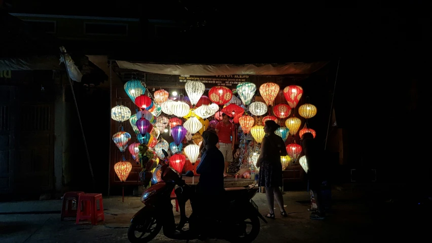 three people standing outside a stall with colorful lanterns on display